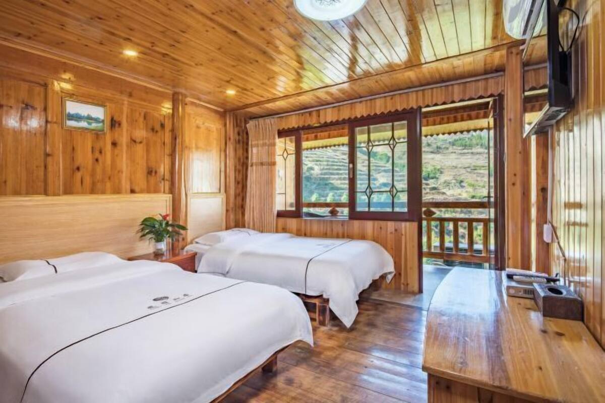 A wooden cabin-style room with two beds, a wooden table, and a large window with a view of a hilly landscape.