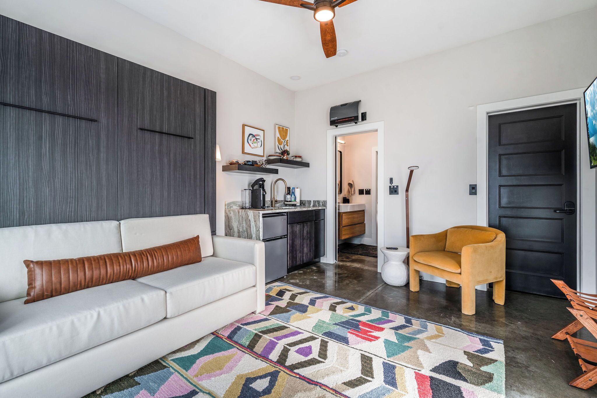A modern living room with a white sofa, a colorful rug, a wooden chair, a kitchen area, and a black door.