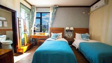 Habitación de hotel con dos camas, un escritorio, una silla, un ventanal con cortinas y vista a la ciudad.