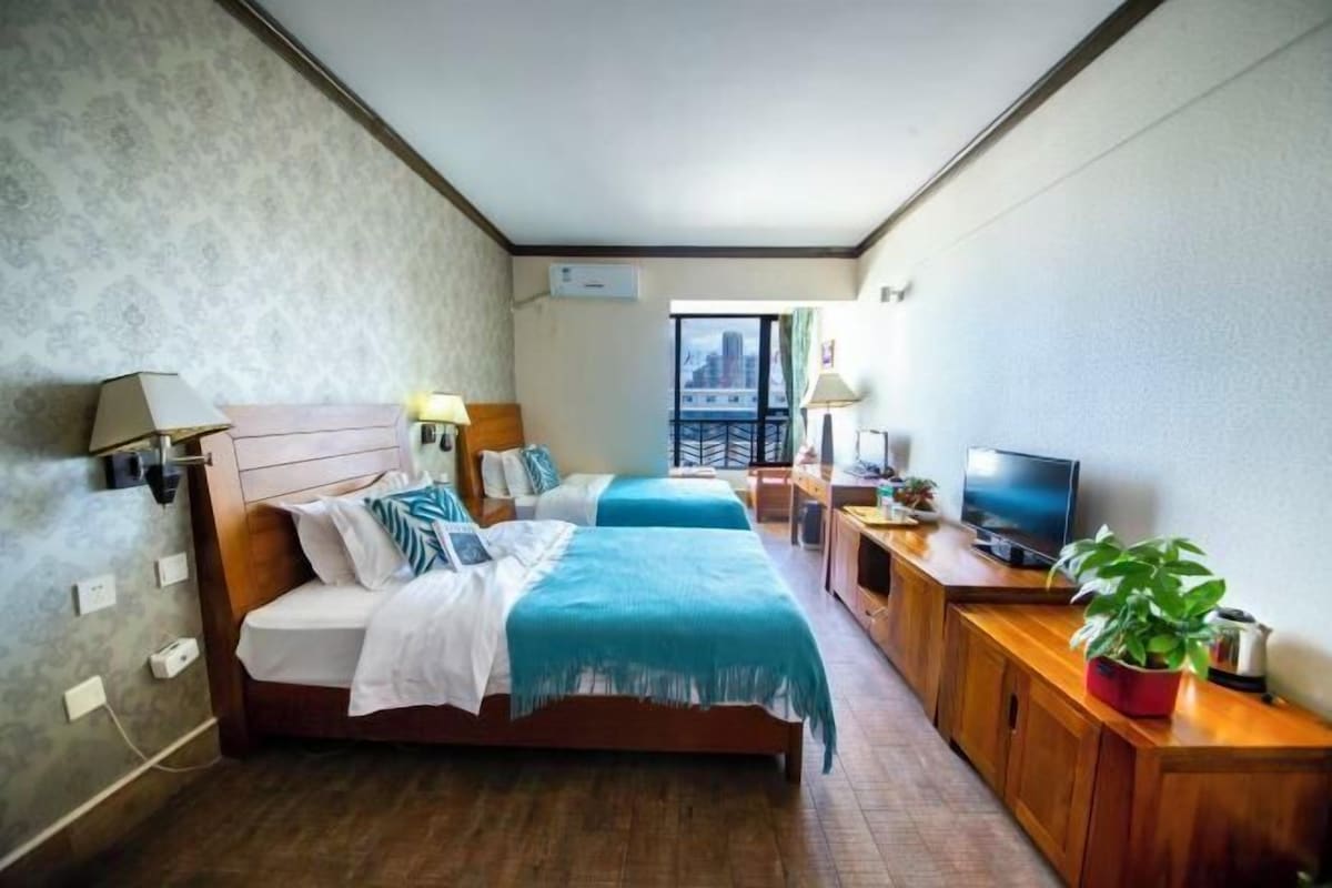 A hotel room with two beds, a TV, a wooden desk, and a balcony with a city view.