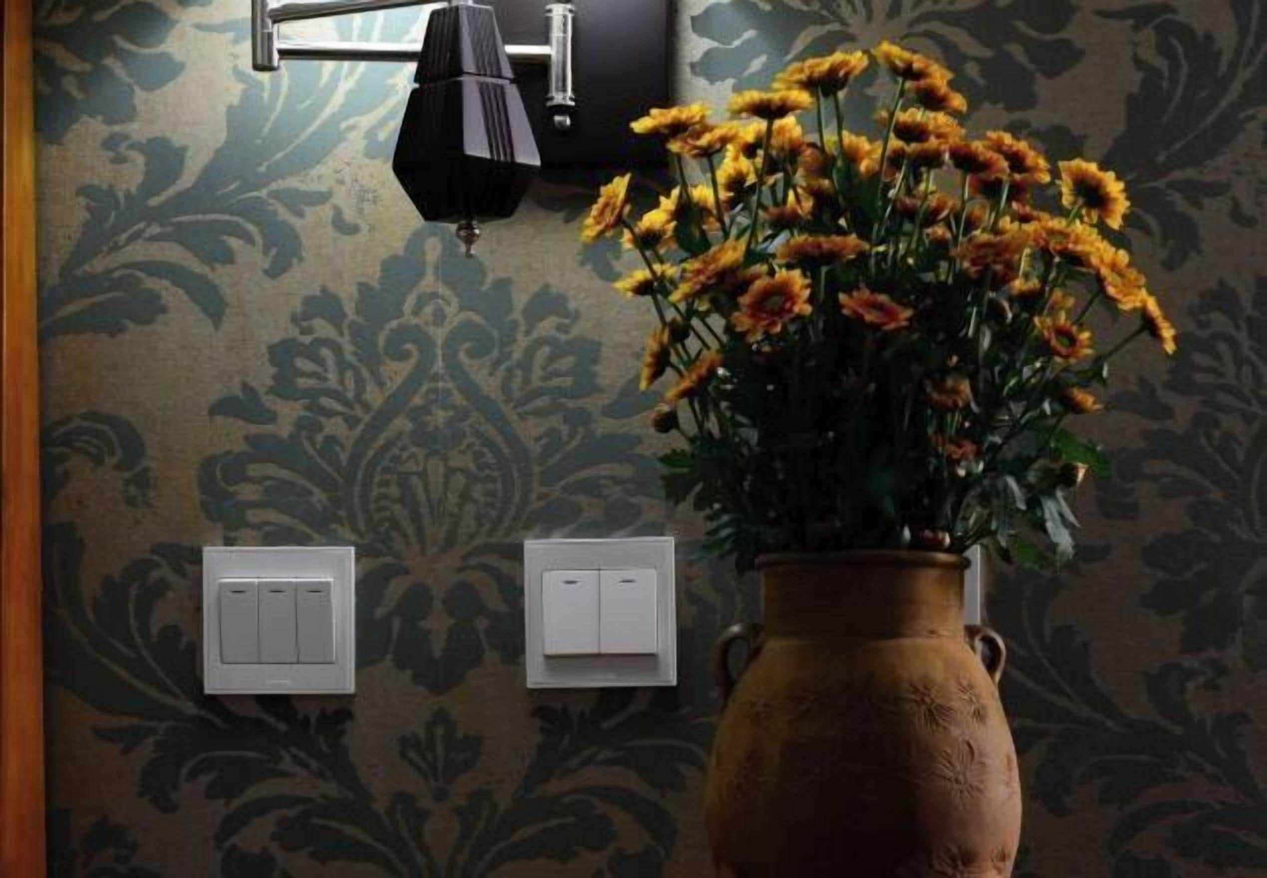 A room with a patterned wallpaper, a vase with yellow flowers, and two wall-mounted light switches.