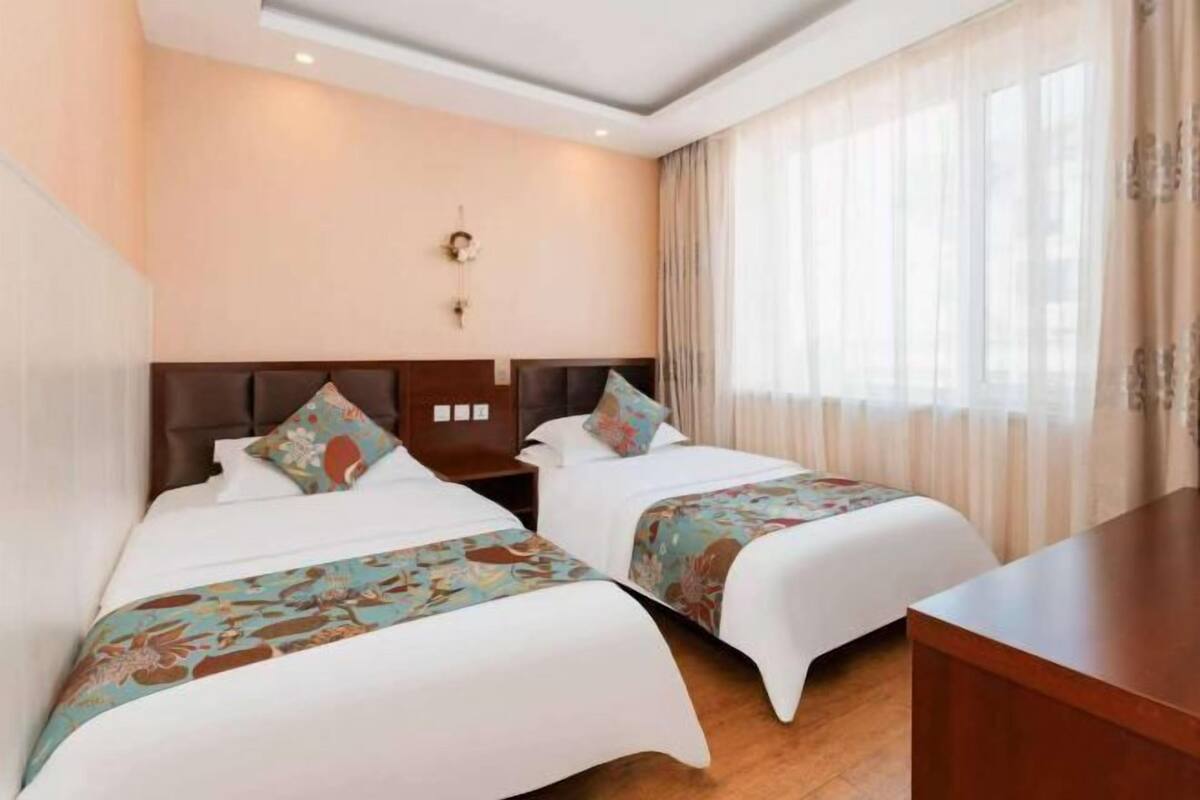 A hotel room with two beds, a wooden nightstand, a desk, and a large window with sheer curtains.