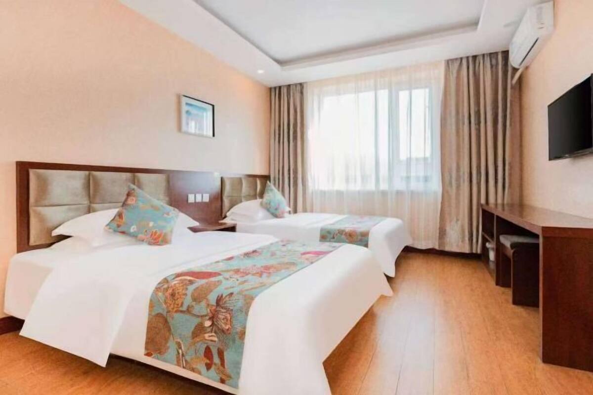 A hotel room with two beds, a TV, a desk, and a large window with curtains.