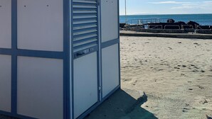En liten, envåningsbyggnad med ett tak av metall och en enda dörr med ett fönster. Den ligger på en sandstrand med en klarblå himmel och en vattenyta i bakgrunden.