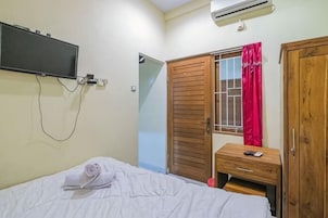 A bedroom with a bed, a TV mounted on the wall, a wooden wardrobe, and a desk with a chair.