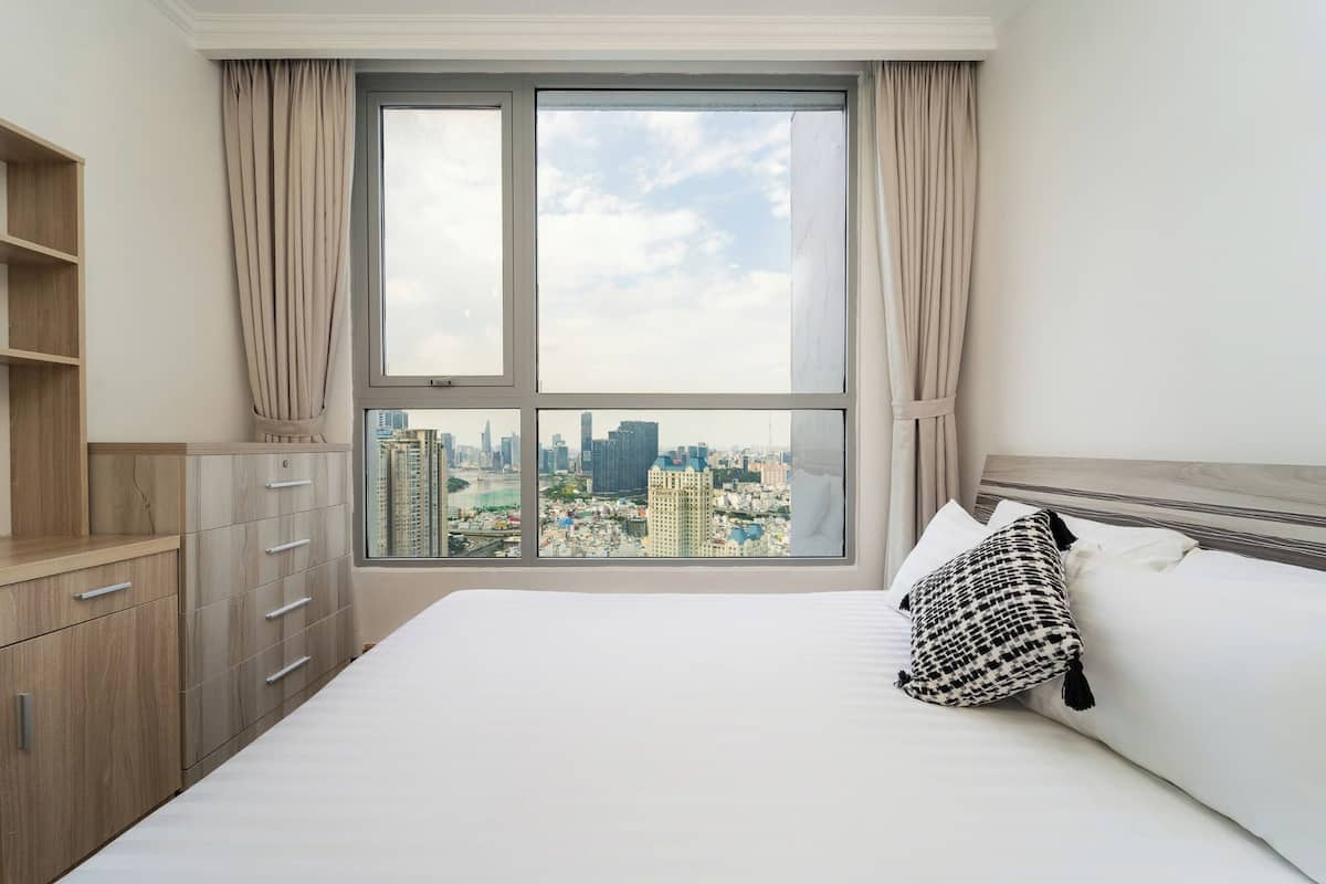 A hotel room with a large window, a bed with white bedding, a black and white pillow, and a wooden wardrobe.