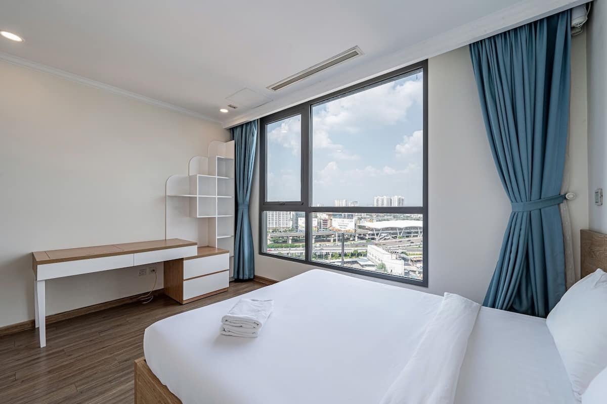A hotel room with a large bed, a desk, and a view of the city through large windows.