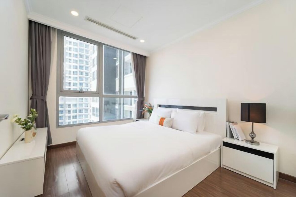 Una habitación de hotel con una cama grande, una mesita de noche con lámpara, una ventana con cortinas y vistas a edificios.