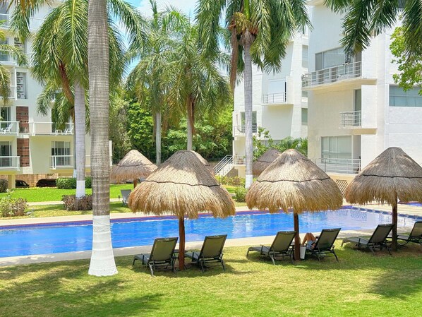 A poolside area with lounge chairs and thatched umbrellas, surrounded by palm trees and a modern building. - Luxury 3 BR Condo in top location near Las Perlas Beach (Cancún)