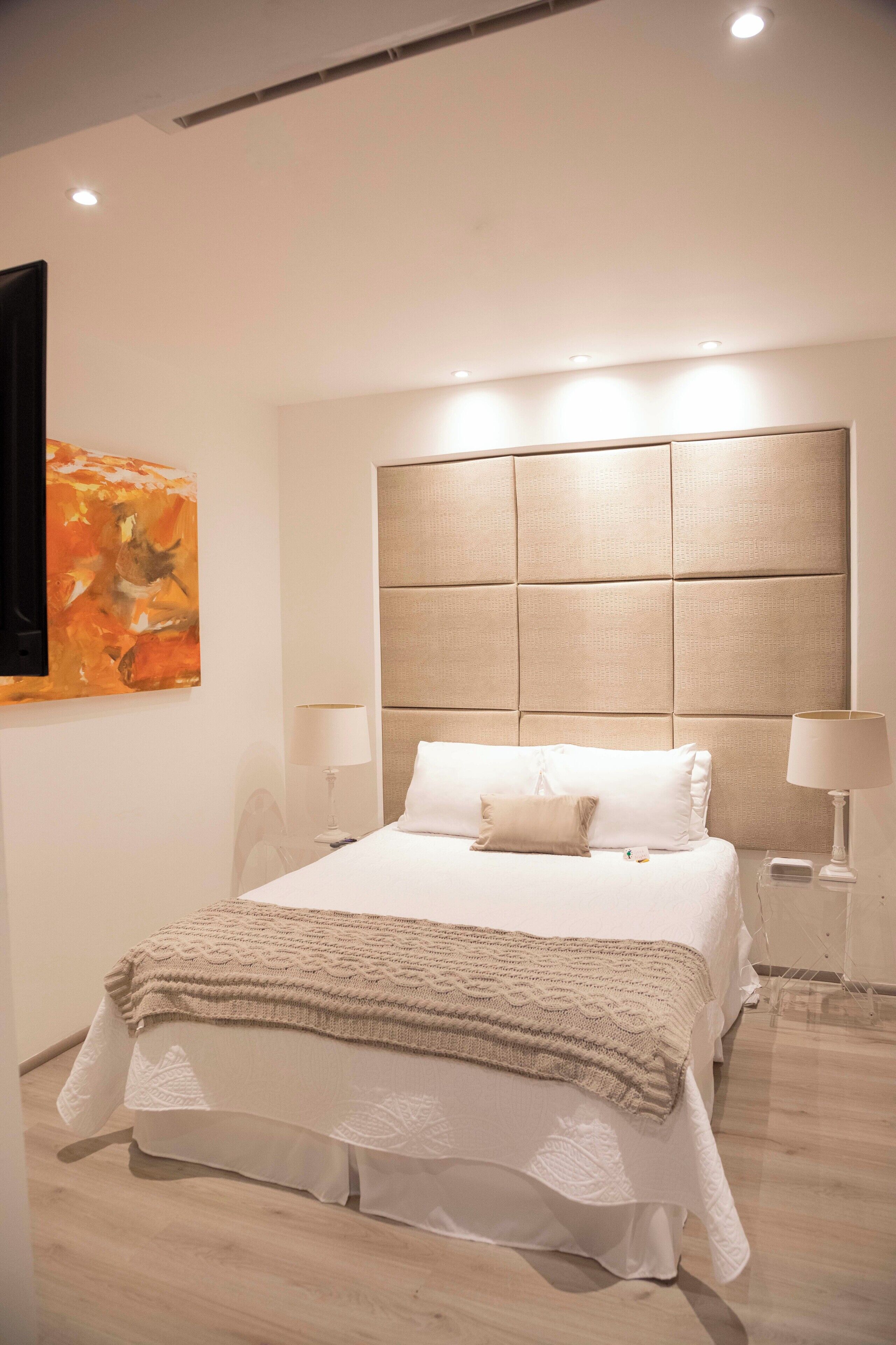 A modern bedroom with a large bed, a headboard with a textured panel, two bedside tables with lamps, and a wall-mounted artwork.