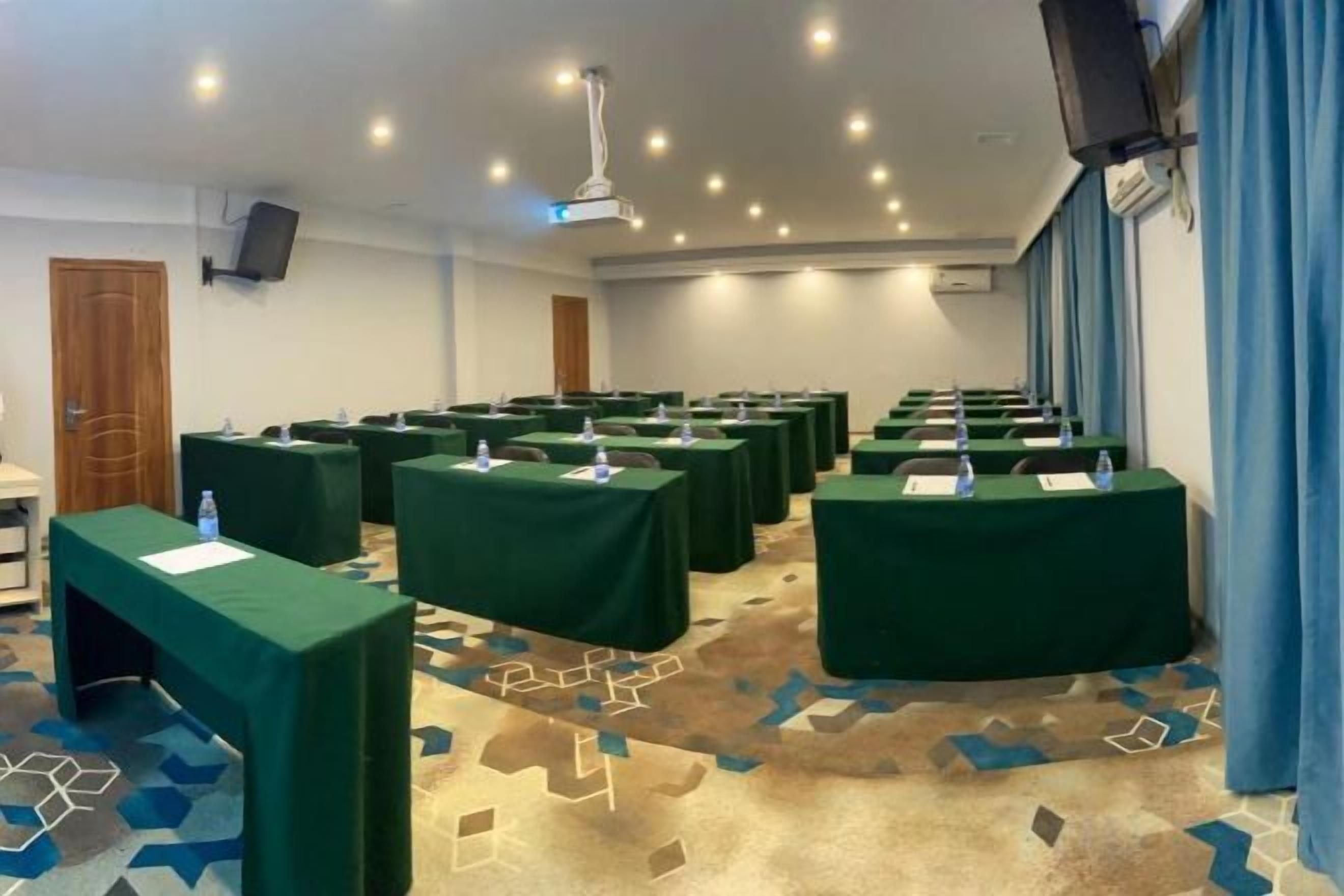 A conference room with multiple tables, each with a green tablecloth and water bottles, in a room with a patterned carpet and blue curtains.