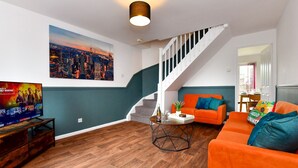 Una sala de estar moderna con un cuadro de un paisaje urbano, una escalera y un televisor mostrando una película.