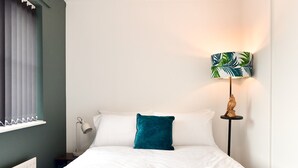 Una cama bien hecha con sábanas blancas y un almohálar turquesa, una mesita de noche con una lámpara y un cuadro colgado en la pared.