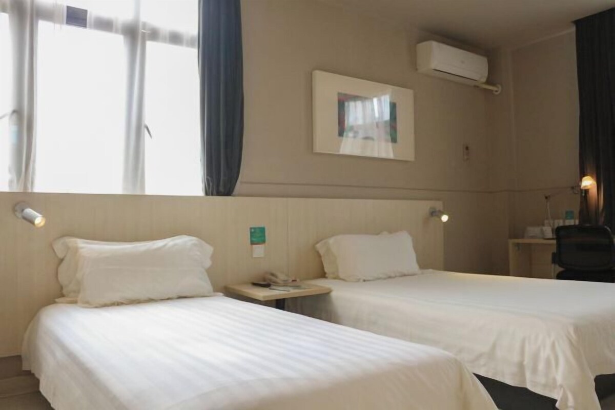 A hotel room with two single beds, a bedside table with a lamp, a small desk with a chair, and a window with curtains.