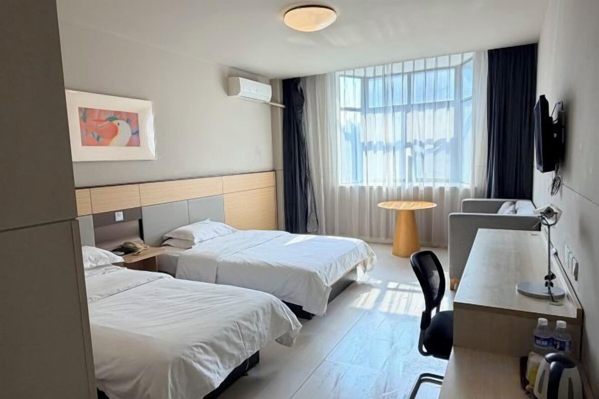 Habitación de hotel con dos camas, un escritorio, una silla, una lámpara y una ventana con cortinas.