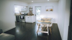 Une cuisine moderne avec des meubles blancs, un coin repas comprenant une table et des chaises, et une porte donnant sur une pièce lumineuse.