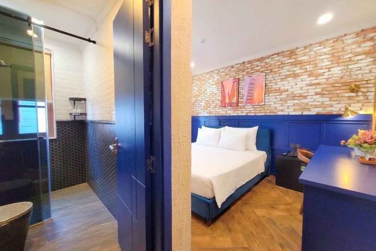 A modern hotel room with a brick-patterned wall, a bed with white bedding, a blue headboard, and a desk with a vase of flowers.
