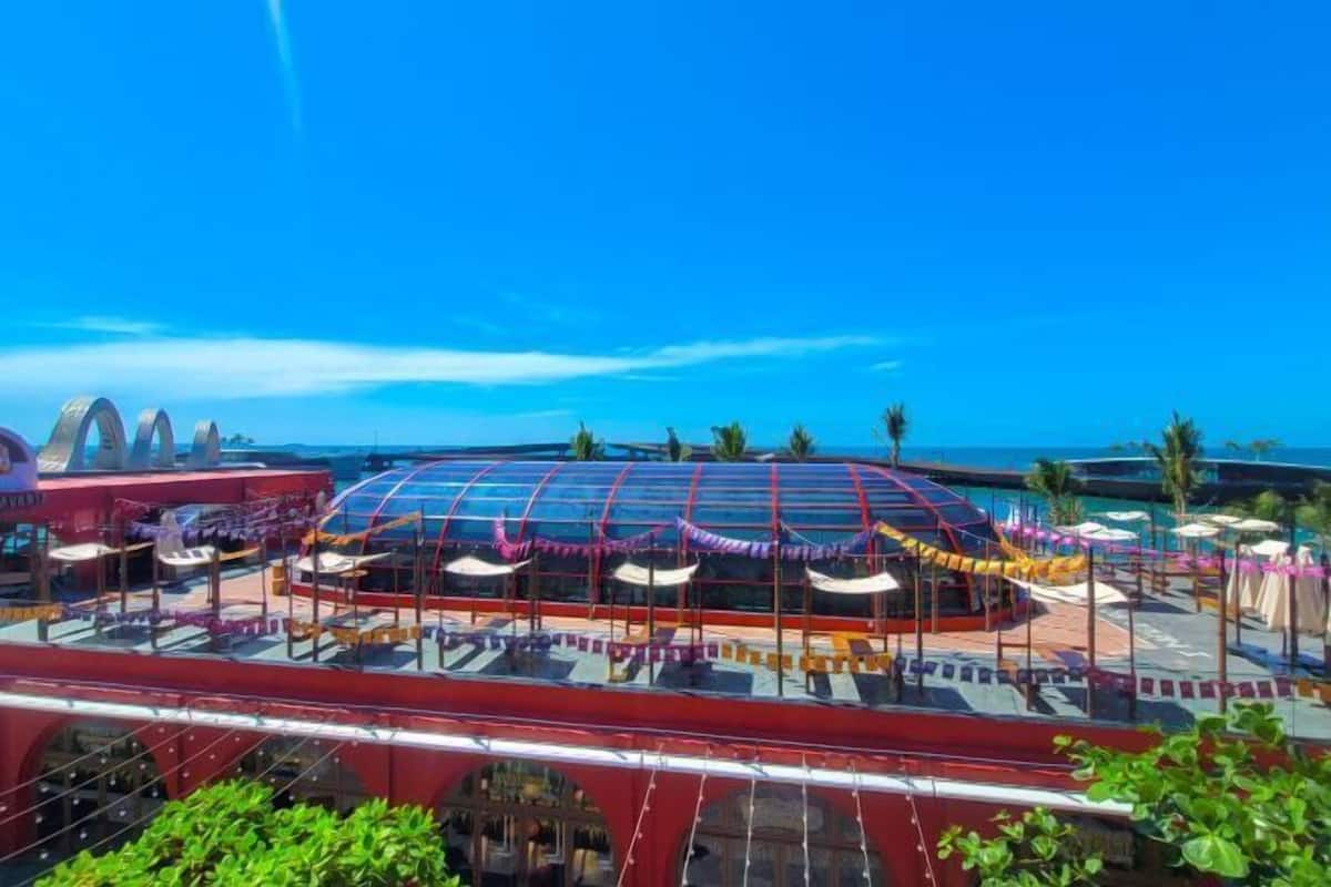 A large, open-air structure with a glass roof, surrounded by palm trees and a clear blue sky.