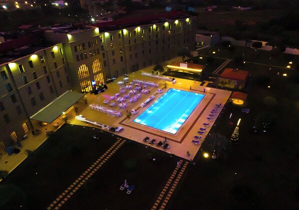 Vue aérienne d’un hôtel doté d’une piscine, d’un espace extérieur aménagé et de plusieurs bâtiments dont les fenêtres sont illuminées.