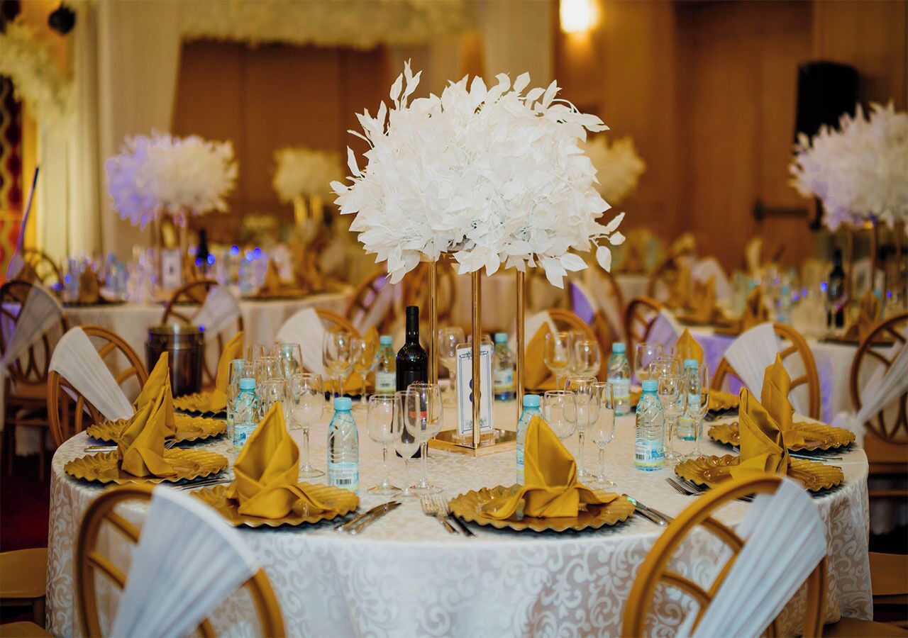 Un salón de banquetes con mesas redondas dispuestas para un evento formal, con manteles blancos, servilletas doradas y centros de mesa florales.