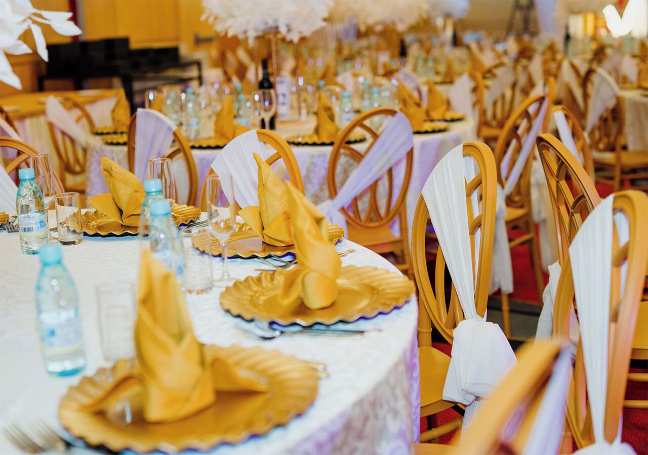 A banquet hall with round tables set for a formal event, each with a white tablecloth, gold napkins, and glassware.