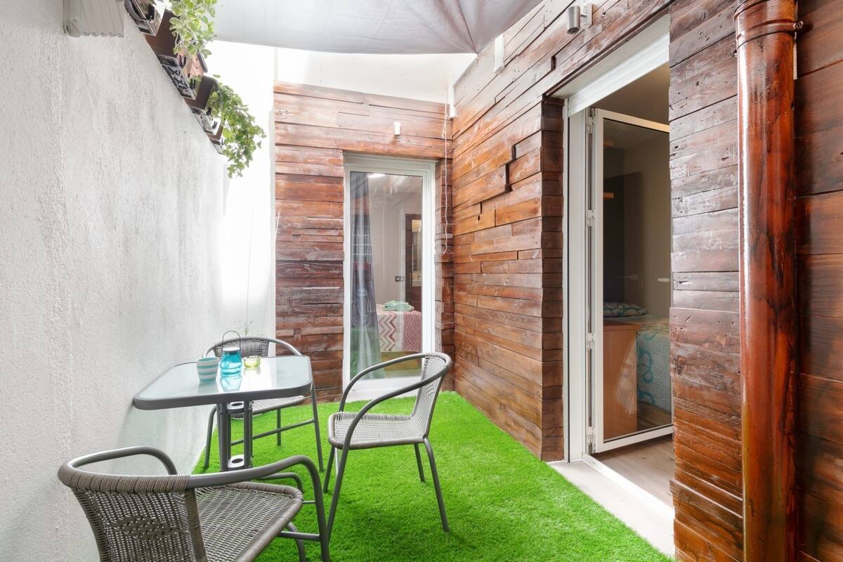 A small patio with a glass table and wicker chairs, a wooden wall, and a sliding glass door leading to an interior room.