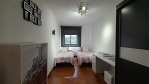 A small, modern bedroom with two beds, a window, and a desk.
