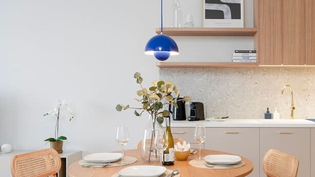 Een moderne keuken met een ronde houten eettafel, vier houten stoelen met beklede zitplaatsen, een blauwe hanglamp en een witte spatwand.