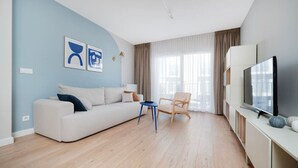 Een moderne woonkamer met een witte bank, een blauwe bijzettafel, een houten stoel, een flatscreen televisie en een groot raam met gordijnen.