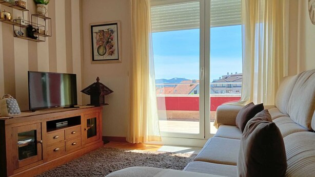 A living room with a sofa, TV, wooden cabinet, and a view of buildings through a glass door. - 3 Bed Flat - Sleeps 6 - Pool - Garden - Parking (Vilagarcia de Arousa)