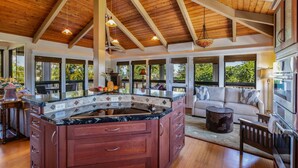 Une cuisine spacieuse dotée d’un grand îlot central, de poutres apparentes en bois au plafond et de grandes fenêtres offrant une vue sur la verdure.