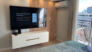 A modern bedroom with a large flat-screen TV mounted on a white cabinet, a wooden slat wall, and a balcony with a city view. - Charming cozy condo in marvelous Honolulu with WiFi (Honolulu)