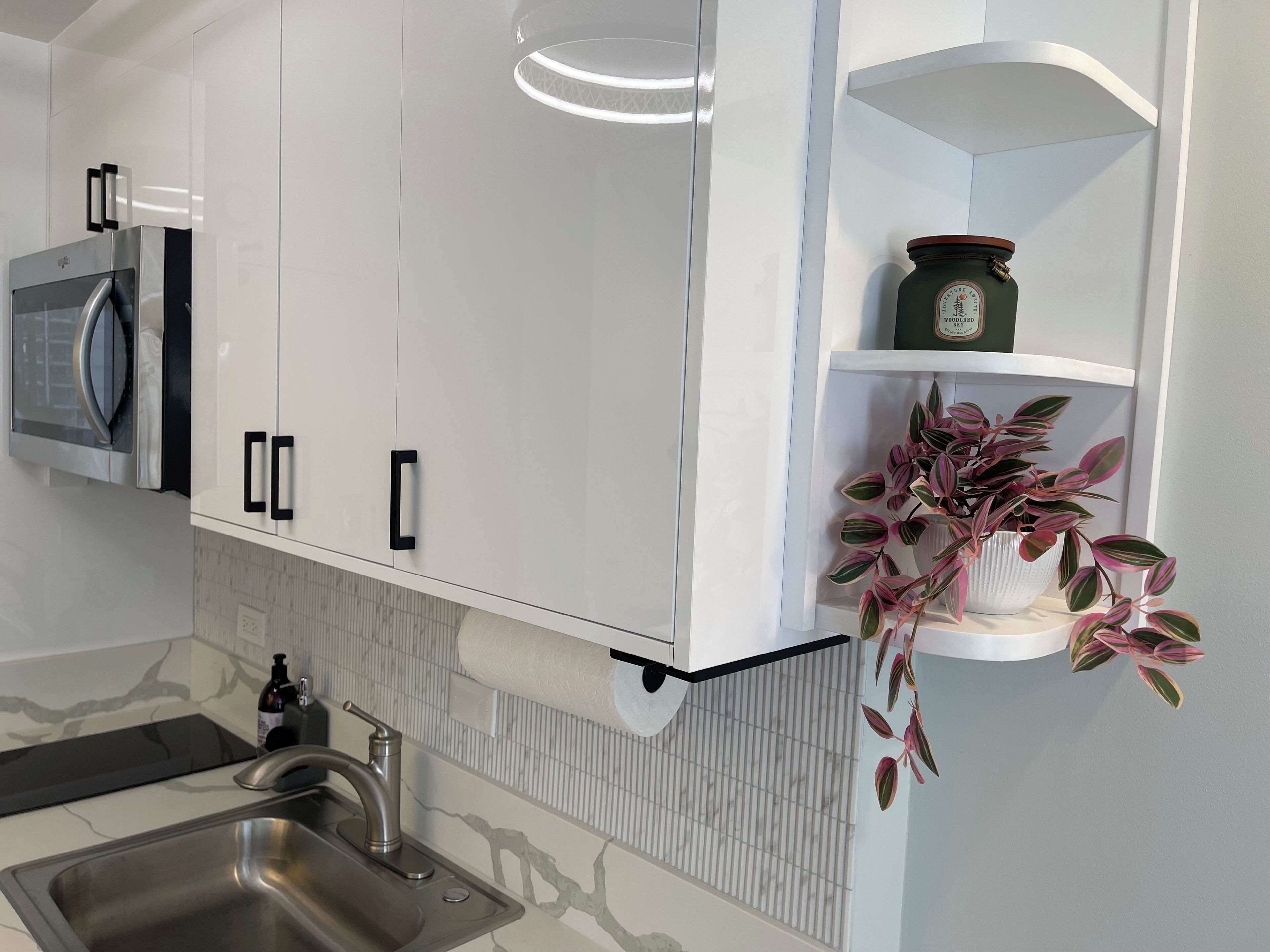 A modern kitchen with white cabinets, a stainless steel sink, a microwave, and a potted plant on a shelf.