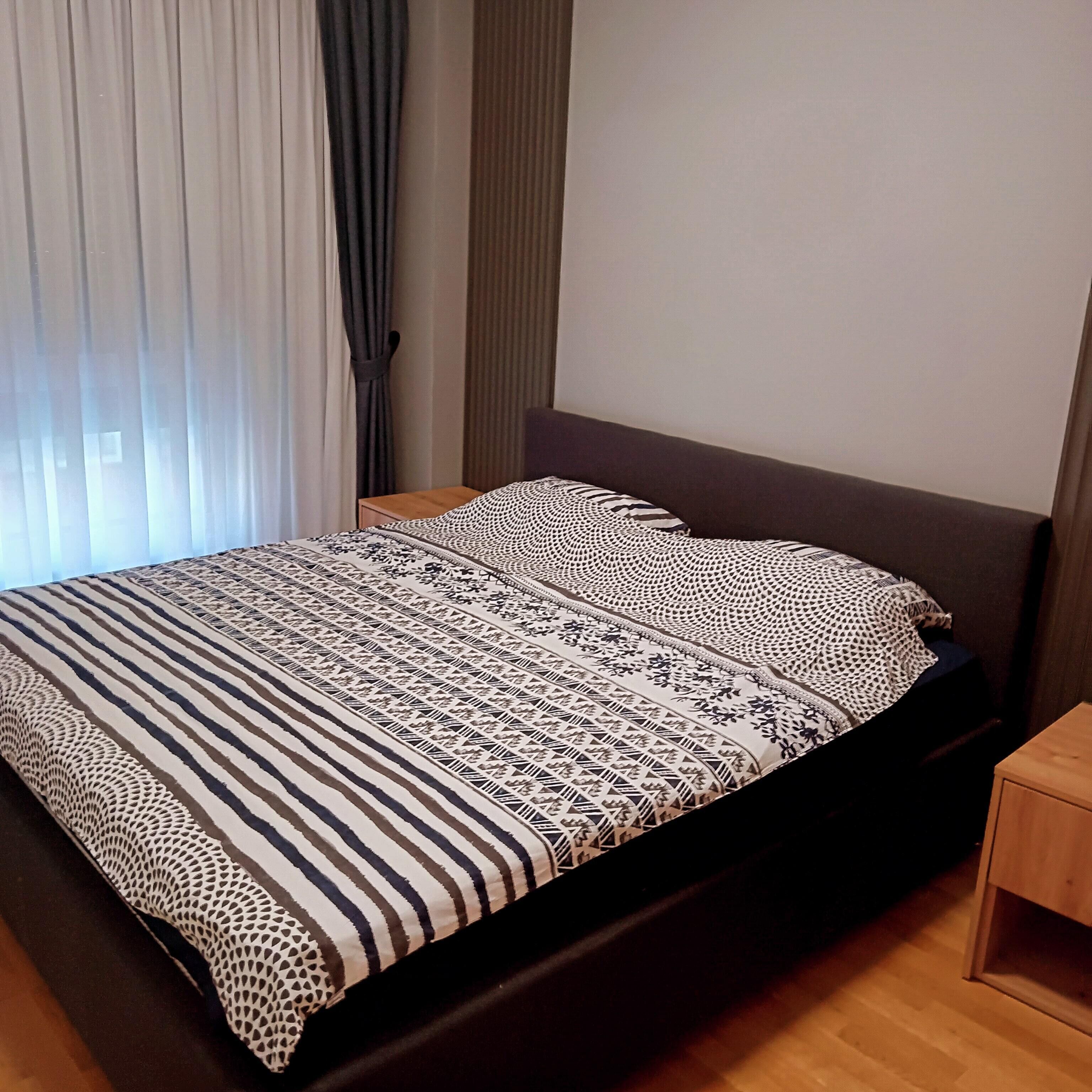 A neatly made bed with a patterned bedspread and pillows, a wooden nightstand, and a window with curtains.