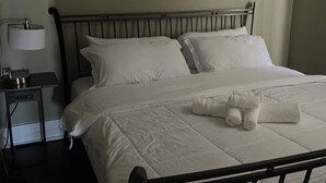 A neatly made bed with white linens and a rolled towel, a bedside table with a lamp, and a painting of a beach scene on the wall. - The L's Luxury 1- Newly renovated Downstairs Apartment (Buffalo)