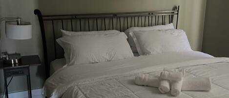 Una cama bien hecha con sábanas blancas y una toalla enrollada, una mesita de noche con una lámpara, y un cuadro de una playa en la pared.