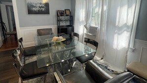 A modern dining area with a glass table and black chairs, a mounted air conditioning unit, a framed inspirational quote, and a window with white curtains.