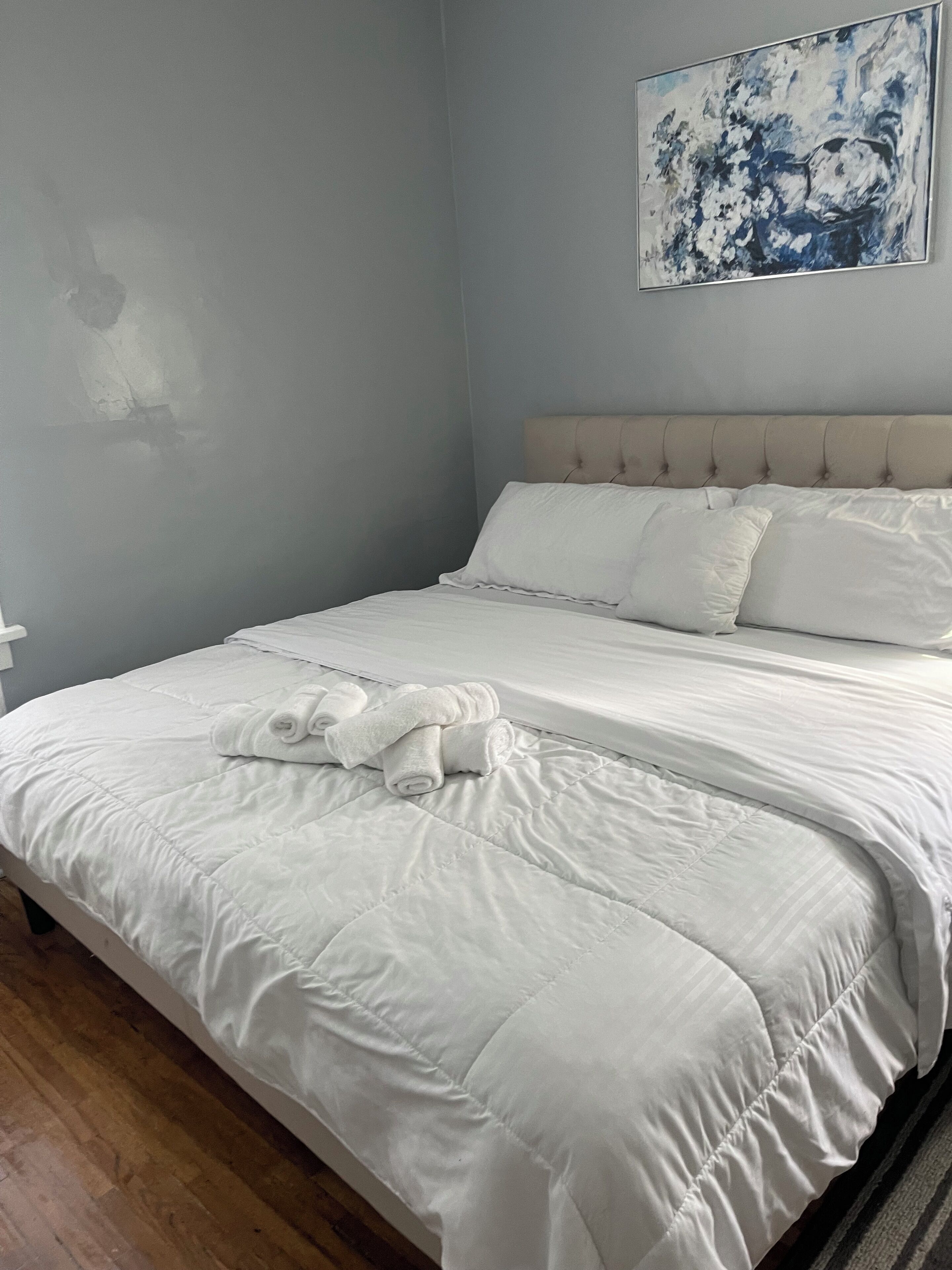 A neatly made bed with white linens and rolled towels, a tufted headboard, and a painting on the wall.
