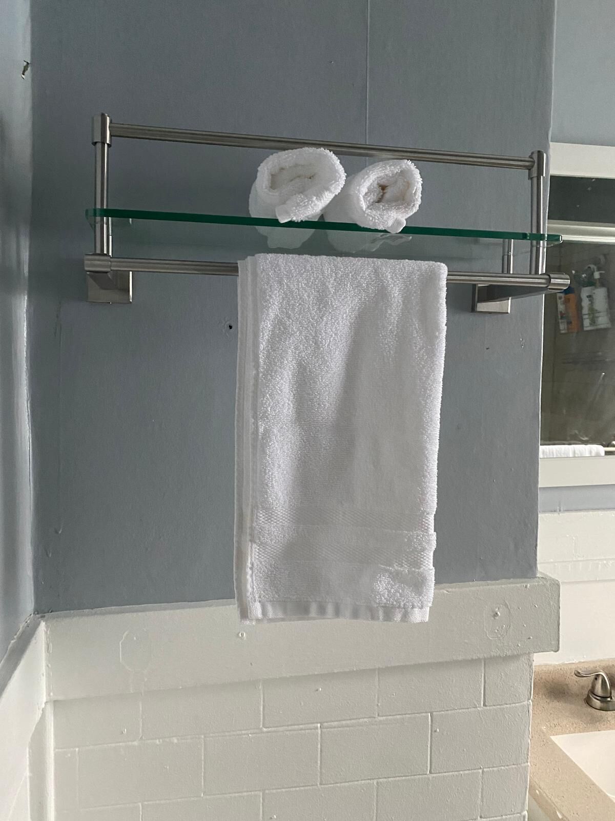 A bathroom with a glass shelf holding two rolled towels and a single towel hanging on a metal towel rack.