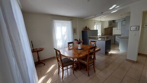 A dining area with a wooden table and chairs, a small side table, and a window with sheer curtains.