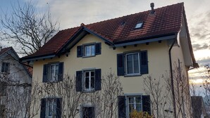 A two-story house with a red-tiled roof, multiple windows with shutters, and a wooden fence in the foreground.