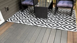 En terrasse med et mønstret tæppe, to fletstole med hynder og et glasbord.