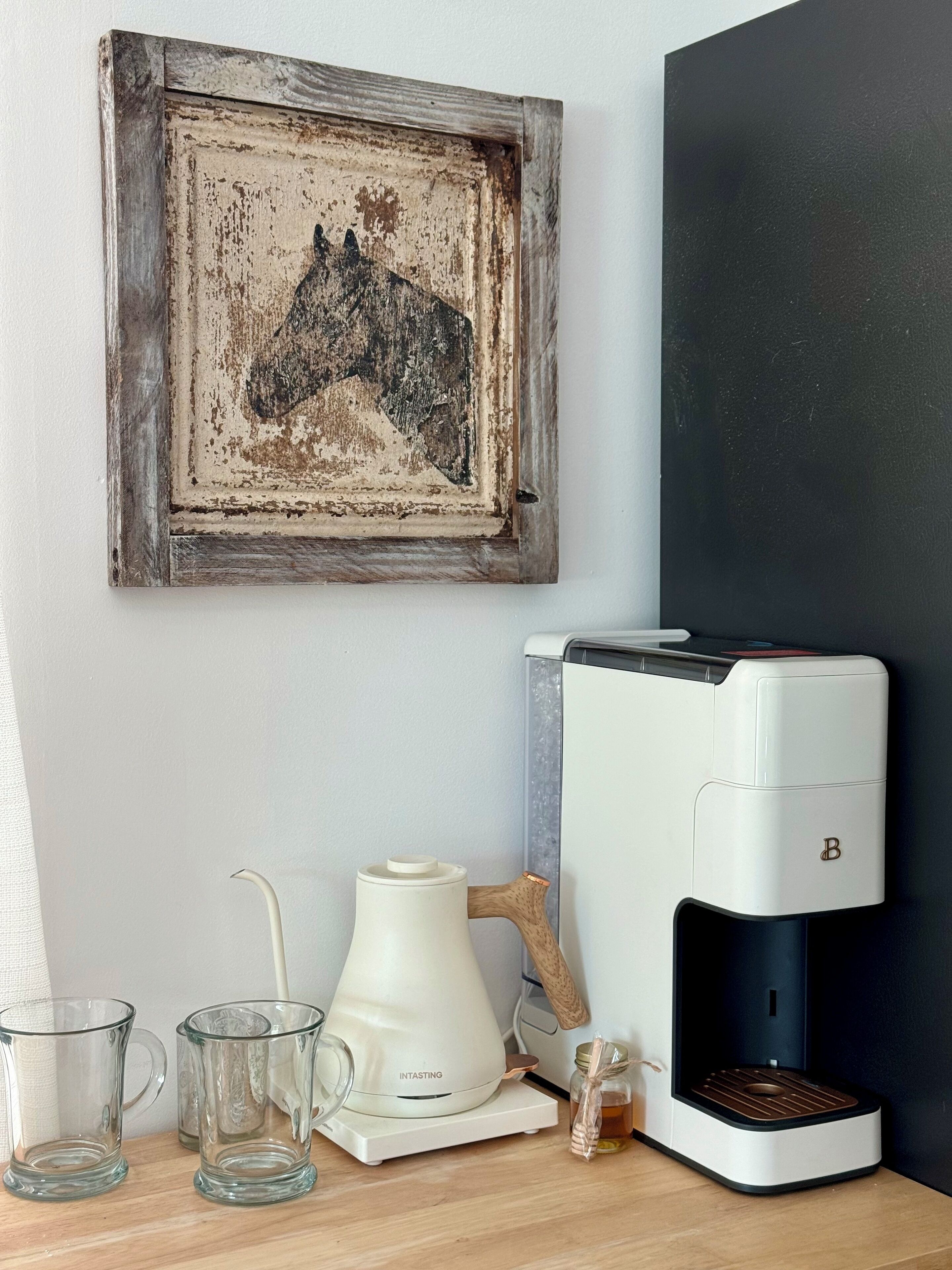 En kaffemaskin, en rustik inramad tavla och två genomskinliga glasmuggar på en träyta.