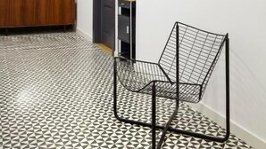Un pasillo con piso de estampado geométrico, un espejo redondo, una puerta oscura, una silla de alambre negra y un cuadro abstracto grande en la pared.