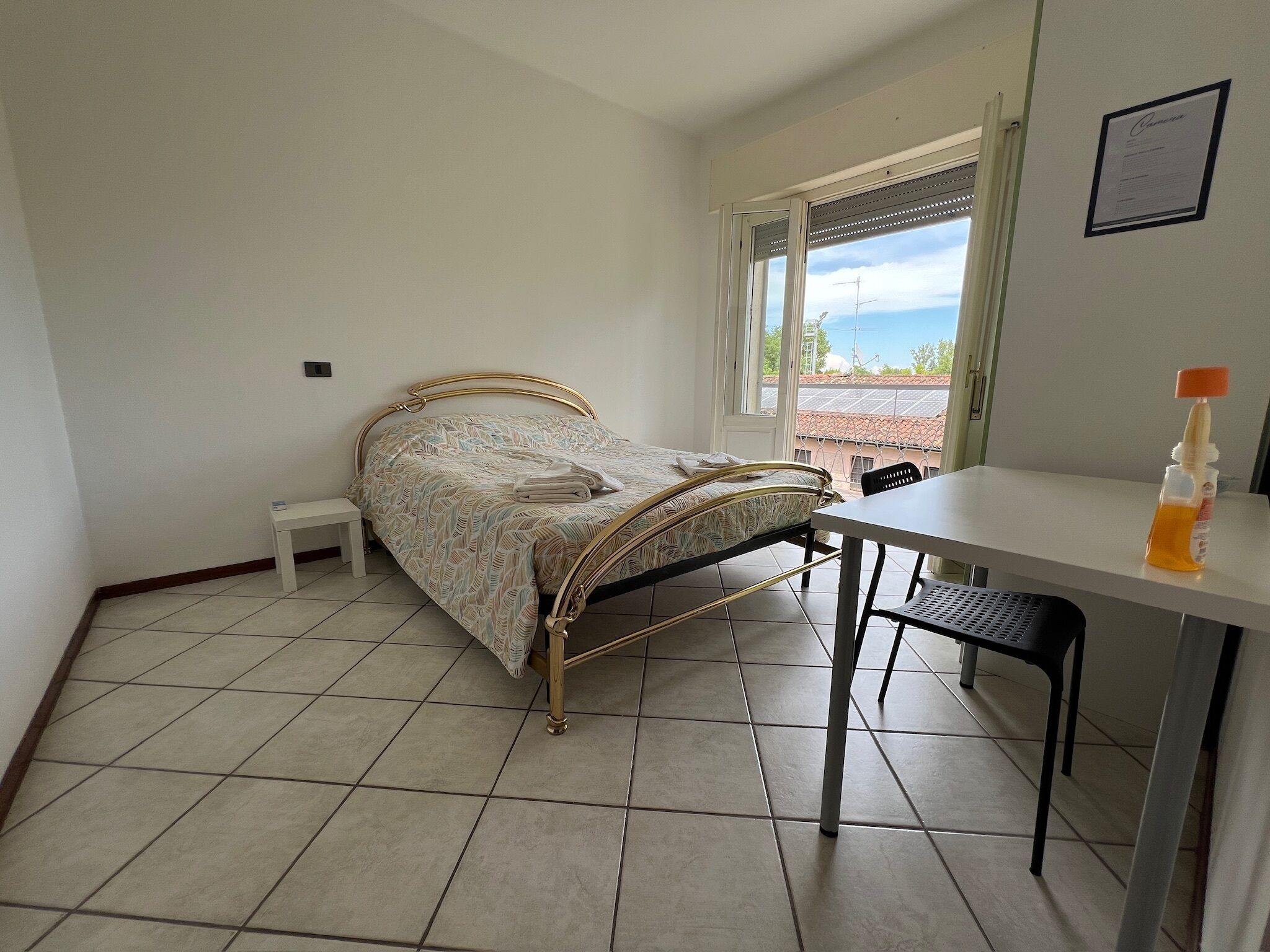 A bedroom with a bed, a small bedside table, a desk with a chair, and a window with a view of the sky and buildings.