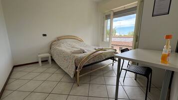 A bedroom with a bed, a small bedside table, a desk with a chair, and a window with a view of the sky and buildings.