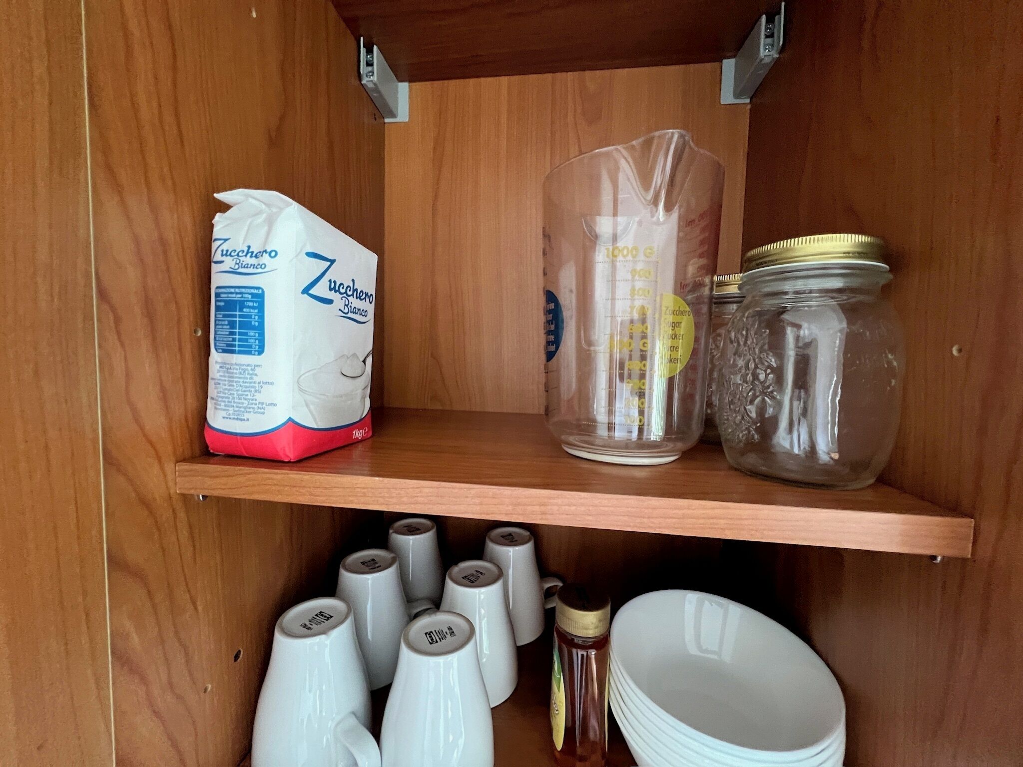 A wooden shelf with a bag of Zucchero Bianco, a measuring cup, a jar, and several white mugs.