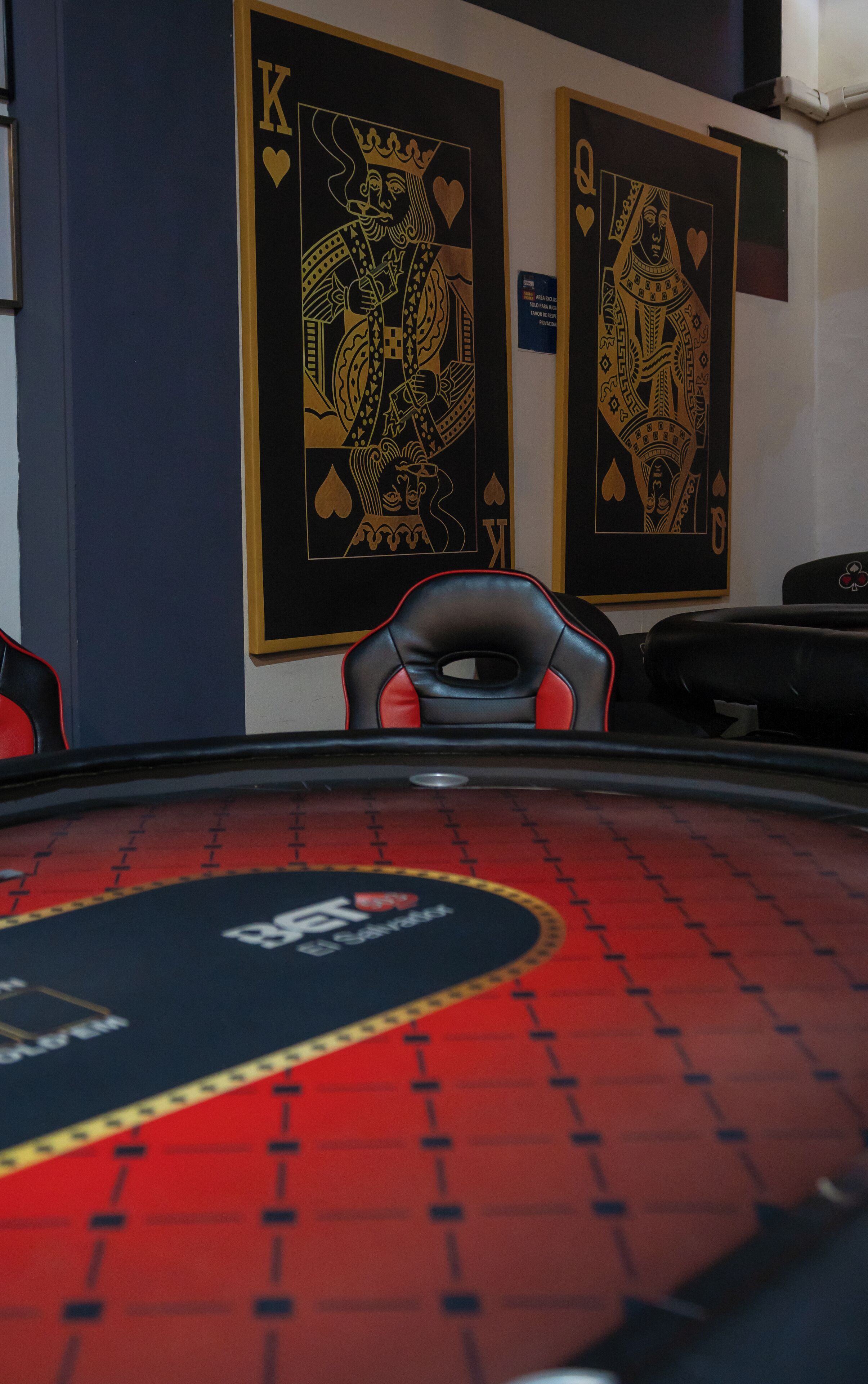 A blackjack table with a red and black design, two playing cards on the wall, and a chair in the foreground.