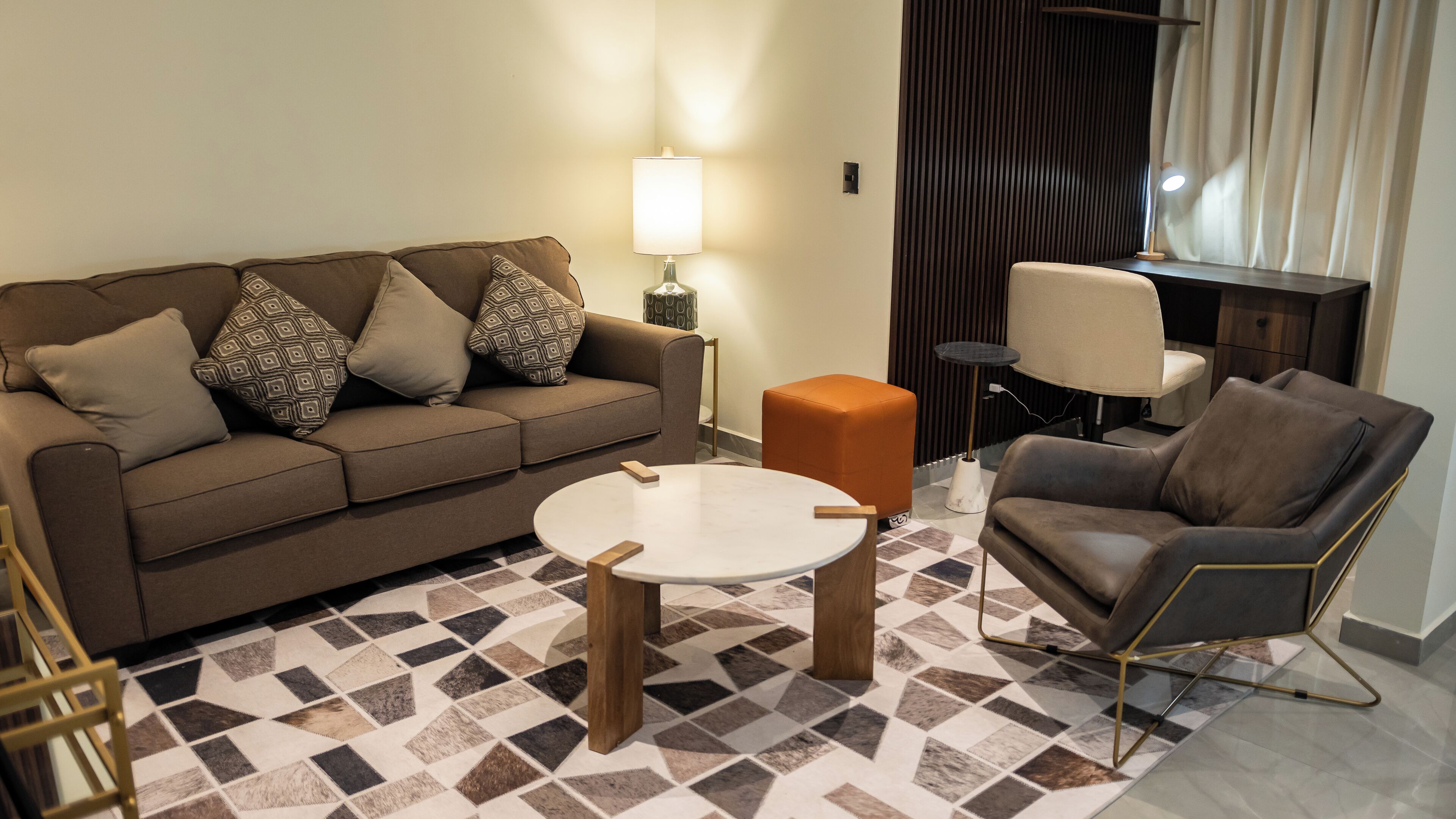 Ein modernes Wohnzimmer mit einem braunen Sofa, einem runden Couchtisch, einem orangefarbenen Hocker und einem Ledersessel.
