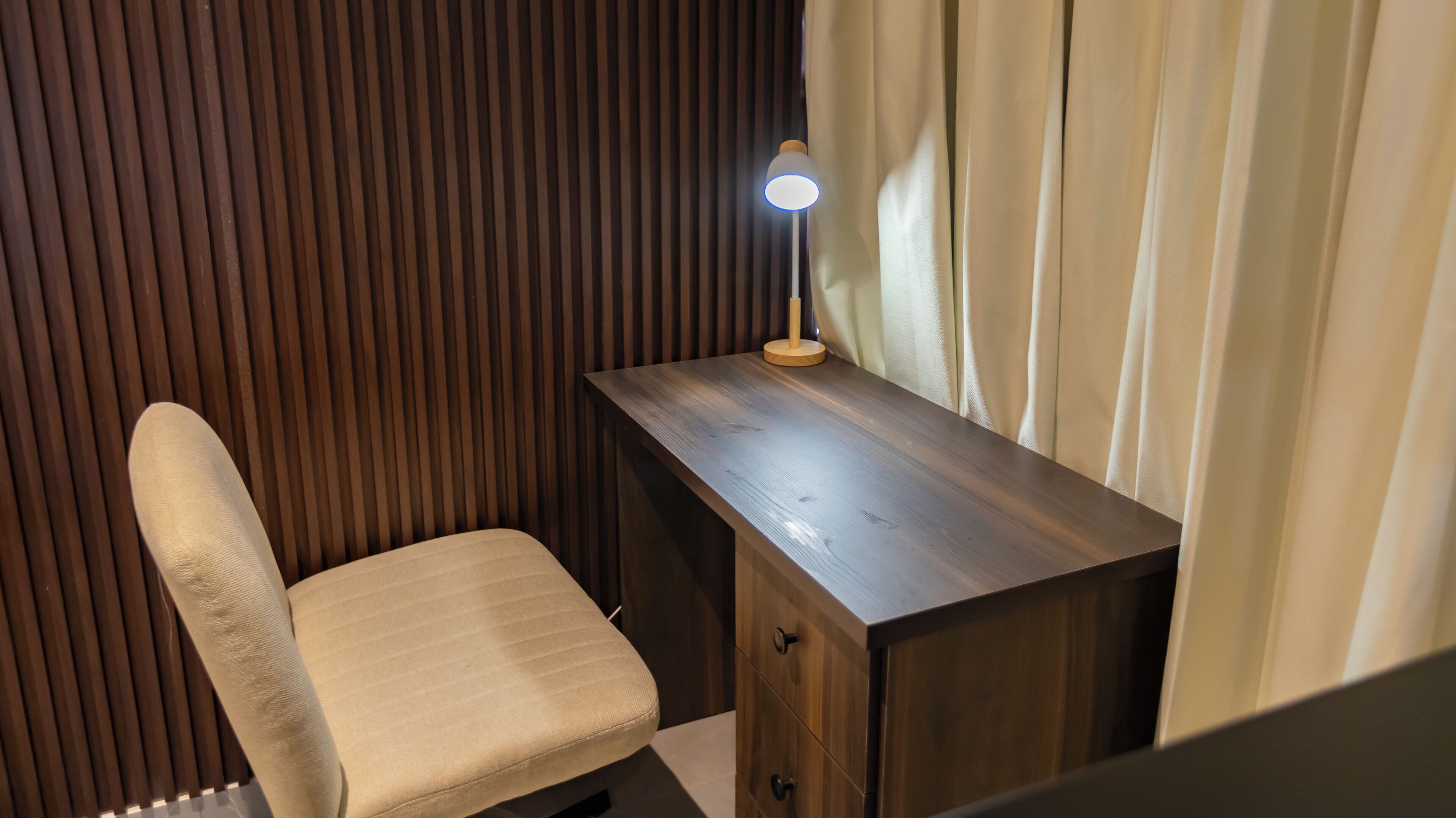 A corner room with a wooden desk, a cushioned chair, and a lamp.